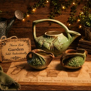 May include: A green ceramic teapot and two matching creamers with a leaf design. The teapot has a large handle and spout. A tag reads "Never Lost Garden Sold Individually". The items are on a wooden surface with a watering can and plants in the background.