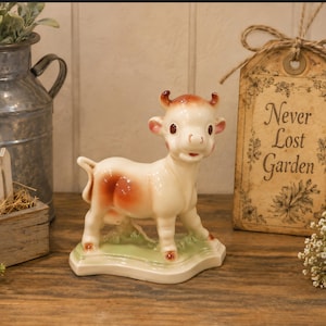 May include: A ceramic cow figurine with a white body and brown spots, horns, and hooves. The cow stands on a green base. A decorative tag with the words "Never Lost Garden" is in the background.