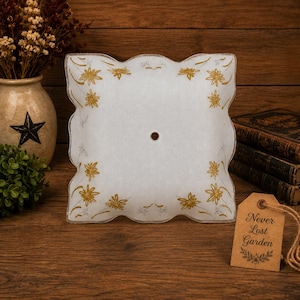 May include: A square, frosted glass light fixture shade with scalloped edges, featuring a gold floral pattern and a central circular opening. A brown paper tag attached with twine displays the text "Never Lost Garden" in black script.