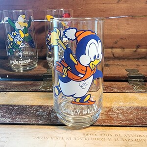 以下が含まれることがあります： 氷の上でスケートをしている漫画のペンギンが描かれた、ビンテージのガラス製の飲み物グラス。ペンギンは青とオレンジの色のマフラーと赤い帽子をかぶっています。グラスの底には「チリー・ウィリー」と書いてあります。