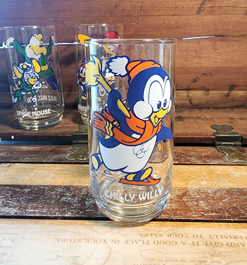 以下が含まれることがあります： 氷の上でスケートをしている漫画のペンギンが描かれた、ビンテージのガラス製の飲み物グラス。ペンギンは青とオレンジの色のマフラーと赤い帽子をかぶっています。グラスの底には「チリー・ウィリー」と書いてあります。