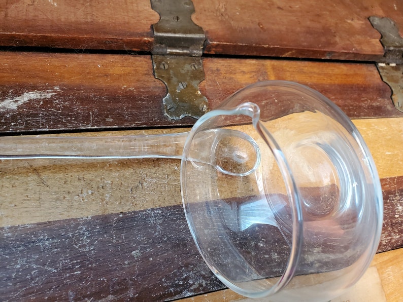 May include: A clear glass ladle with a long handle. The ladle is resting on a wooden surface.