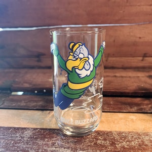 以下が含まれることがあります： 透明なガラス製の飲みグラス。緑、黄色、青の鳥の漫画が描かれており、くちばしは白く、羽根も白いです。鳥は飛んでおり、グラスの底には「Buzz Buzzard」という文字が印刷されています。