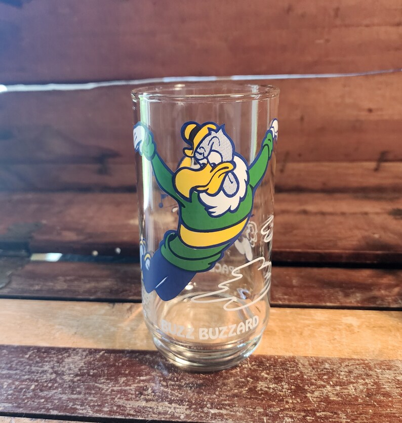 以下が含まれることがあります： 透明なガラス製の飲みグラス。緑、黄色、青の鳥の漫画が描かれており、くちばしは白く、羽根も白いです。鳥は飛んでおり、グラスの底には「Buzz Buzzard」という文字が印刷されています。