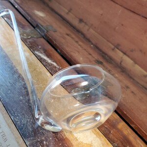May include: A clear glass ladle with a long handle, resting on a wooden surface.