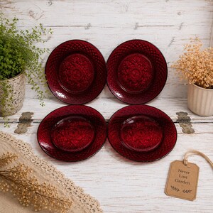 May include: Four circular plates made of deep red pressed glass, each featuring a detailed embossed floral pattern in the center and a textured diamond grid design along the rim. The plates are arranged in a two-by-two grid on a white surface.