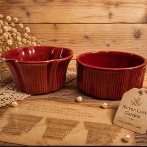 May include: Two burgundy-colored ceramic bowls with vertical ribbed detailing. One bowl has a scalloped rim, the other is round. A tag reads "Never Lost Garden" and "sold individually". The bowls sit on a wooden surface.