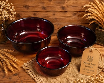 May include: Three ruby red glass bowls of varying sizes. The bowls have a dark rim and a glossy finish. A tag reads "Never Lost Garden sold individually". The bowls are on a wooden surface.