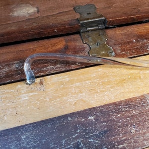 May include: A clear glass curved rod with a rounded end, lying on a wooden surface with a light brown stain.