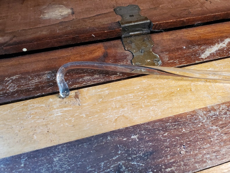 May include: A clear glass curved rod with a rounded end, lying on a wooden surface with a light brown stain.