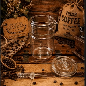 May include: A clear glass coffee carafe with a spout, lid, and a glass stirring rod. A burlap bag of fresh coffee beans is behind the carafe. The bag reads "FRESH COFFEE 100% ARABICA NET WT. 1 LB."