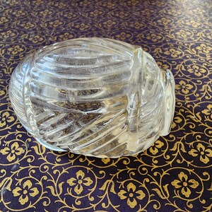 May include: A clear, molded glass paperweight shaped like a rounded, ribbed shell or yarn ball. The object features a series of raised, diagonal ridges that wrap around its curved surface, creating a textured, translucent appearance throughout.
