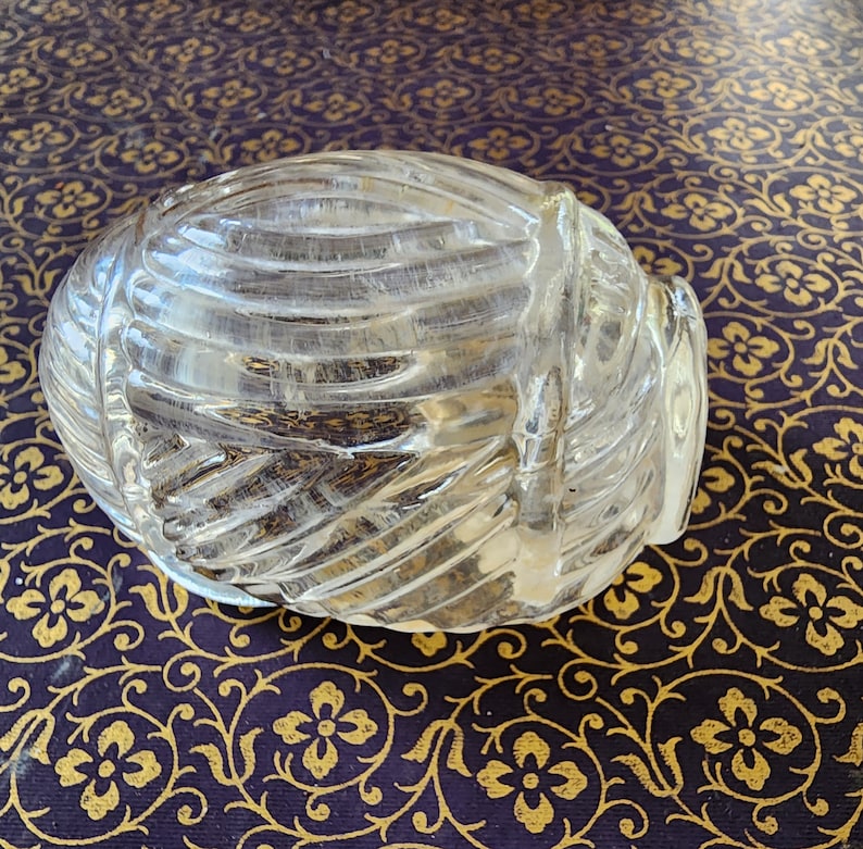 May include: A clear, molded glass paperweight shaped like a rounded, ribbed shell or yarn ball. The object features a series of raised, diagonal ridges that wrap around its curved surface, creating a textured, translucent appearance throughout.