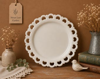 May include: A white ceramic plate with a decorative openwork heart-shaped rim. The plate is displayed alongside a small ceramic bird figurine, a metal pitcher containing dried flowers, a small vase with dried stems, and a hanging tag with the text Never Lost Garden.