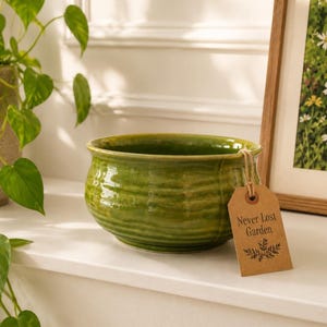May include: A green ceramic bowl with a textured, ribbed design. The bowl has a wide rim and a tag that reads "Never Lost Garden". The bowl is sitting on a white surface, with a plant and a framed picture in the background.