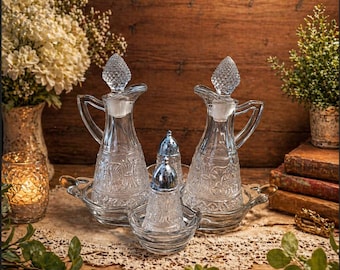 May include: A clear glass cruet set with two decorative bottles, a salt and pepper shaker, and a small bowl. The bottles have a handle and a stopper. The set is on a glass tray.