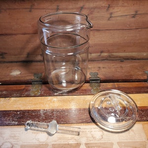 May include: A clear glass jar with a lid and a glass pouring spout. The jar has a textured surface and is sitting on a wooden surface.