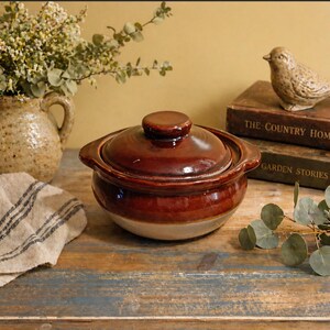 May include: A brown ceramic casserole dish with a lid, featuring a two-tone design. The dish has a rounded shape with handles and a small knob on the lid. The background includes a bird figurine and vintage books.