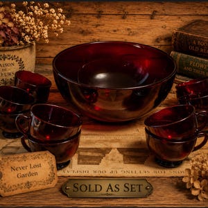 May include: A set of ruby red glass serving bowls and cups. The set includes a large bowl, a smaller bowl, and several cups. The cups are stacked in pairs. The set is displayed on a wooden surface with vintage books and dried flowers.