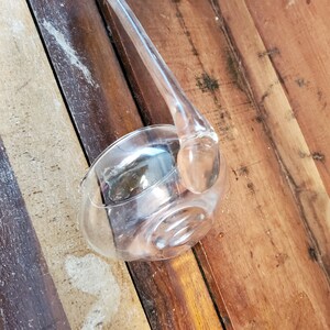 May include: A clear glass ladle with a long handle. The ladle is resting on a wooden surface.