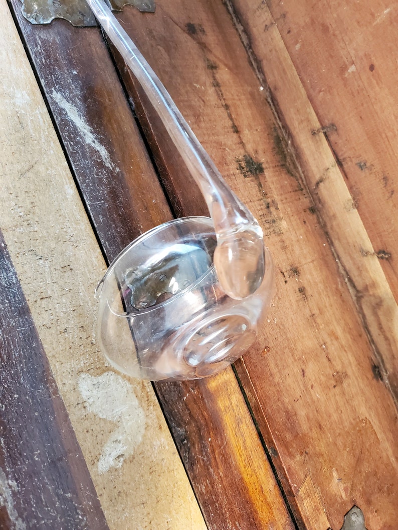 May include: A clear glass ladle with a long handle. The ladle is resting on a wooden surface.