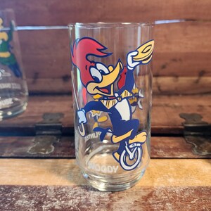 以下が含まれることがあります： ウッディー・ウッドペッカーがユニサイクルに乗っている漫画の画像が描かれた透明なガラス製の飲み物グラス。グラスの底には「ウッディー」と印刷されています。