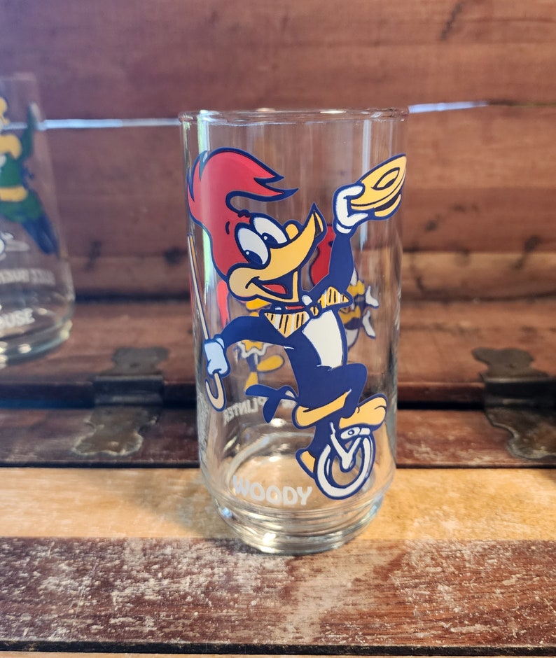 以下が含まれることがあります： ウッディー・ウッドペッカーがユニサイクルに乗っている漫画の画像が描かれた透明なガラス製の飲み物グラス。グラスの底には「ウッディー」と印刷されています。