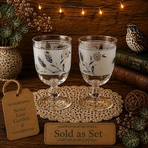 May include: Two clear glass goblets featuring a frosted band with a black and gold floral design. Each glass has a short, rounded stem and a wide base. A brown tag reads Never Lost Garden and a rectangular sign reads Sold as Set.