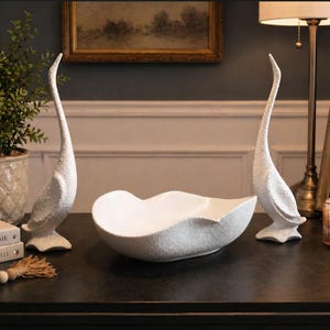 May include: White textured ceramic swan figurines and a matching bowl on a dark surface. The swans have long necks and stand on small bases. The bowl has a unique, wavy shape. Decorative home decor.