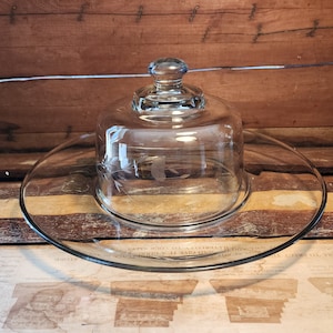 May include: A clear glass cheese dome with a decorative etched design sits on a matching clear glass plate. The dome has a knob on top.
