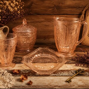 May include: A still life arrangement featuring vintage pink glassware, including a pitcher, a covered jar, a tumbler, and a decorative dish. A decorative tag with the text "Never Lost Garden" hangs in the background, along with a vintage key.