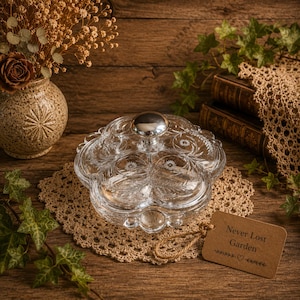 May include: A clear, multi-compartment glass serving dish with a flower-shaped design and a central silver-toned knob handle. The glass features etched floral and leaf patterns on each section. A brown paper tag with the text Never Lost Garden is attached.