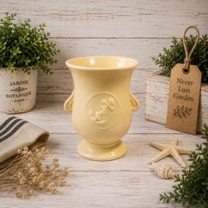 May include: A pale yellow ceramic vase with a flared rim and a pedestal base. The vase features two small side handles and a circular embossed design on the front depicting a nautical anchor surrounded by a rope-like border detail.