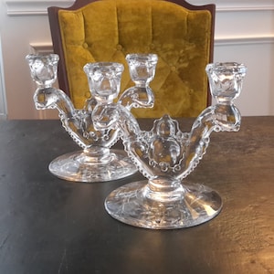 May include: Two clear glass candle holders with ornate designs and three candle cups each. The candle holders are on a dark wooden surface.