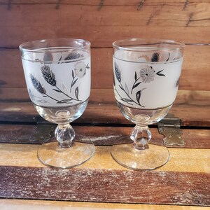 May include: Two clear glass goblets with frosted white bases and silver trim. Each goblet features a black and white floral design with a silver band around the middle.