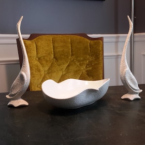 May include: Two white ceramic bird sculptures and a white ceramic bowl with a textured surface. The bowl is sitting on a dark brown table. The birds are standing upright with their necks arched.