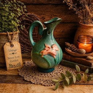 May include: A teal-colored vintage pitcher with a coral-colored floral design. The pitcher sits on a crocheted doily, with a lit candle and a tag that reads "Vintage Never Lost Garden". The background is a rustic wooden surface.