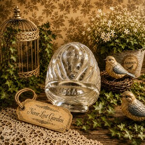 May include: A clear glass egg-shaped object with vertical ribbing and the letters U.S.A. embossed on the front, accompanied by a rectangular tan tag with the text Never Lost Garden, positioned near a gold wire birdcage and two bird figurines.