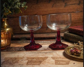 Könnte beinhalten: Zwei klare Weingläser mit rubinroten Stielen und Böden. Die Gläser stehen auf einer Holzoberfläche, die einen Vintage-Look erzeugt. Eine goldfarbene Vase und ein Kerzenhalter sind im Hintergrund zu sehen.
