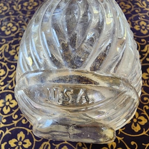 May include: A clear, molded glass paperweight shaped like an egg with a textured, ribbed surface pattern. The glass features the letters U.S.A. embossed in a horizontal band across the lower section of the object, with a smooth, rounded top.