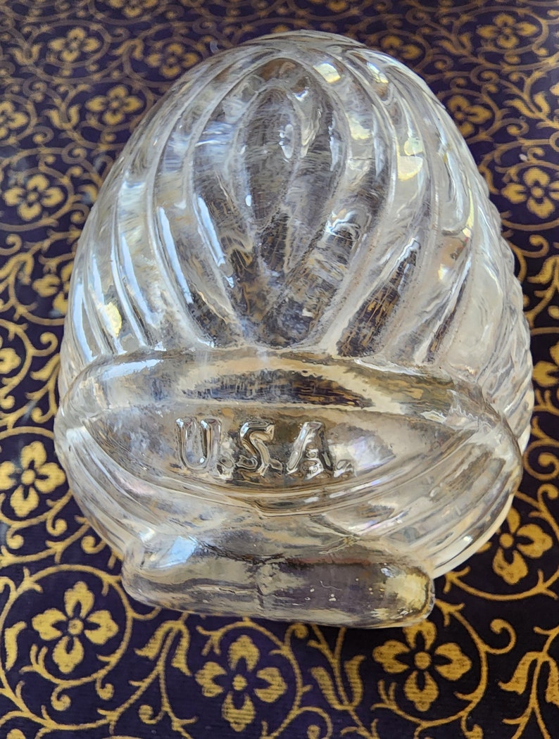 May include: A clear, molded glass paperweight shaped like an egg with a textured, ribbed surface pattern. The glass features the letters U.S.A. embossed in a horizontal band across the lower section of the object, with a smooth, rounded top.
