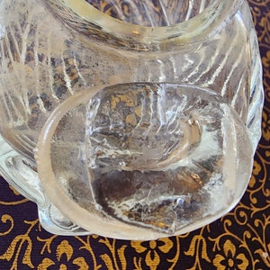 May include: A clear glass container featuring a textured, ribbed surface pattern and a thick, rounded glass lid. The lid has a smooth, slightly irregular top surface and sits partially offset on the opening of the container, revealing the interior.