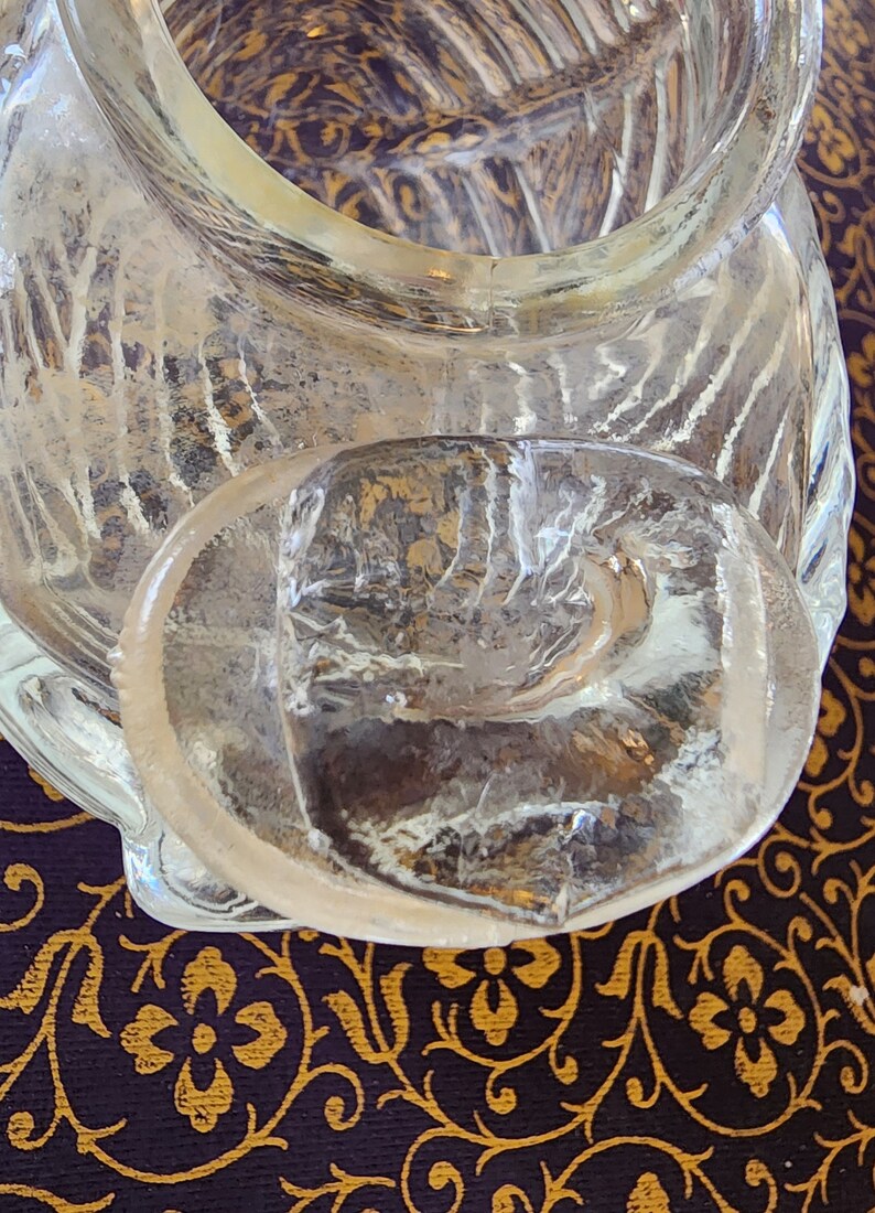 May include: A clear glass container featuring a textured, ribbed surface pattern and a thick, rounded glass lid. The lid has a smooth, slightly irregular top surface and sits partially offset on the opening of the container, revealing the interior.