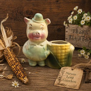 May include: A ceramic planter shaped like a pig wearing overalls standing next to a yellow corn cob. The pig has a light yellow body with pink cheeks and a small hat. The planter is designed with a hollow section for plants within the corn cob.