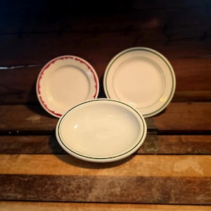 May include: Three white ceramic plates with decorative rims. One plate has a red scalloped edge, another has a green border, and the third is an oval serving dish with a green border. The plates are arranged on a wooden surface.