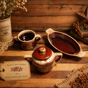 May include: A rustic still life featuring a brown ceramic sugar bowl with a lid, a small mug, and an oval baking dish. The items are arranged on a wooden surface with a tag that reads "Never Lost Garden" and "Sold Individually."