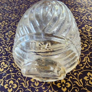 May include: A clear, molded glass object with a rounded, ribbed, and textured surface. The glass features a prominent horizontal band across the middle with the letters U.S.A. embossed in a raised, sans-serif font. The overall shape is bulbous.