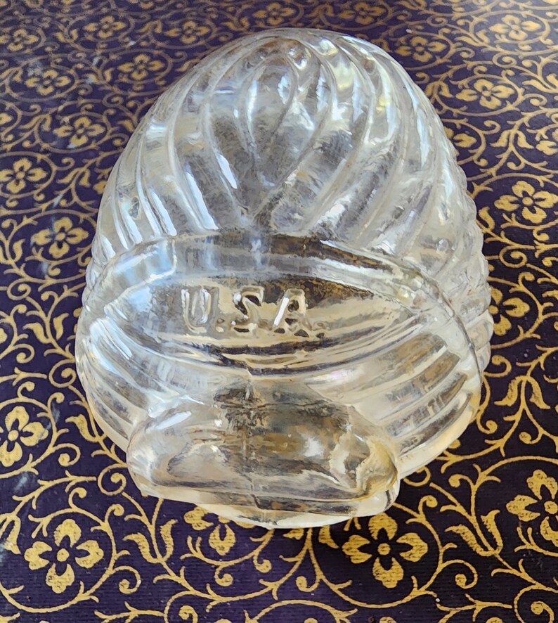 May include: A clear, molded glass object with a rounded, ribbed, and textured surface. The glass features a prominent horizontal band across the middle with the letters U.S.A. embossed in a raised, sans-serif font. The overall shape is bulbous.
