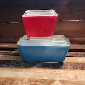 May include: Two vintage Pyrex square storage containers with glass lids. One is a vibrant red, stacked atop a blue container. The containers are rectangular with slightly flared sides. The lids are clear glass. The containers are on a wooden surface.
