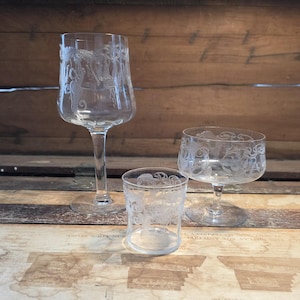 May include: Three clear glass drinking glasses with etched floral designs. The glasses include a tall wine glass, a champagne coupe, and a small tumbler. The glasses are arranged on a wooden surface.
